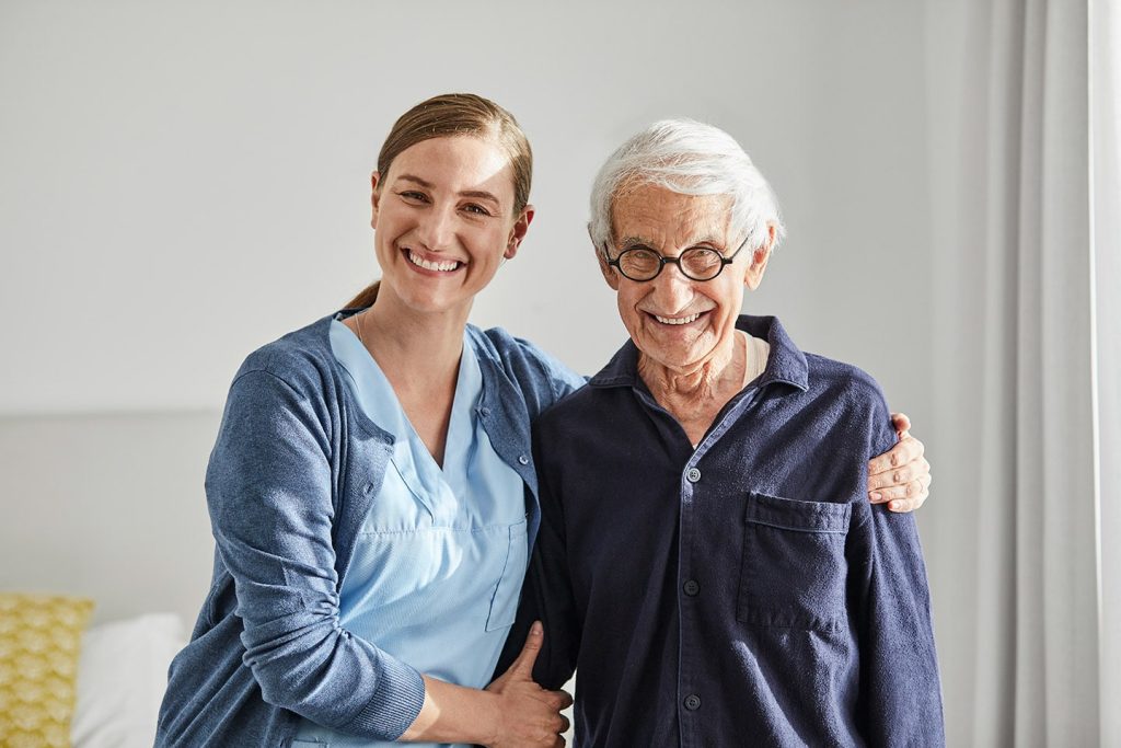 A nurse standing arm in arm with a smiling older man in his room