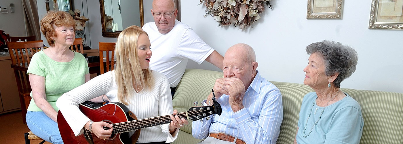 A music therapist sings and plays guitar beside an older man playing a harmonica, with three family members listening in a living room