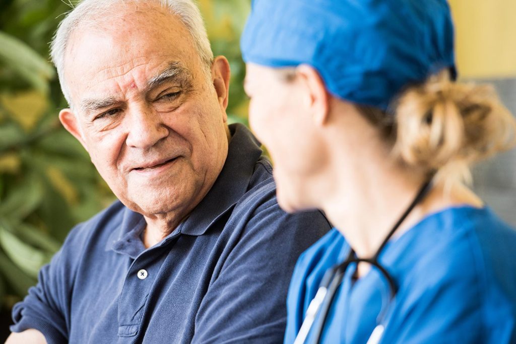 An older man in a navy polo shirt listening to a nurse in blue scrubs and surgical cap