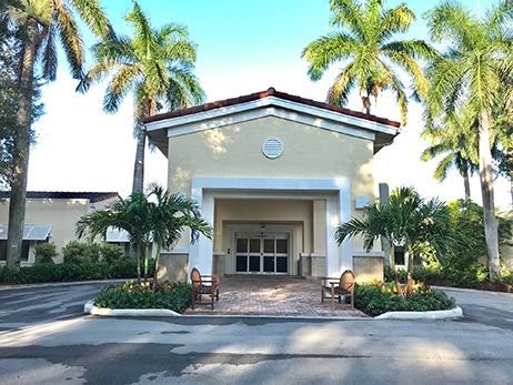 The palm-lined entrance portico of the Care Center at Boca Raton, with benches either side of the brick-paved driveway