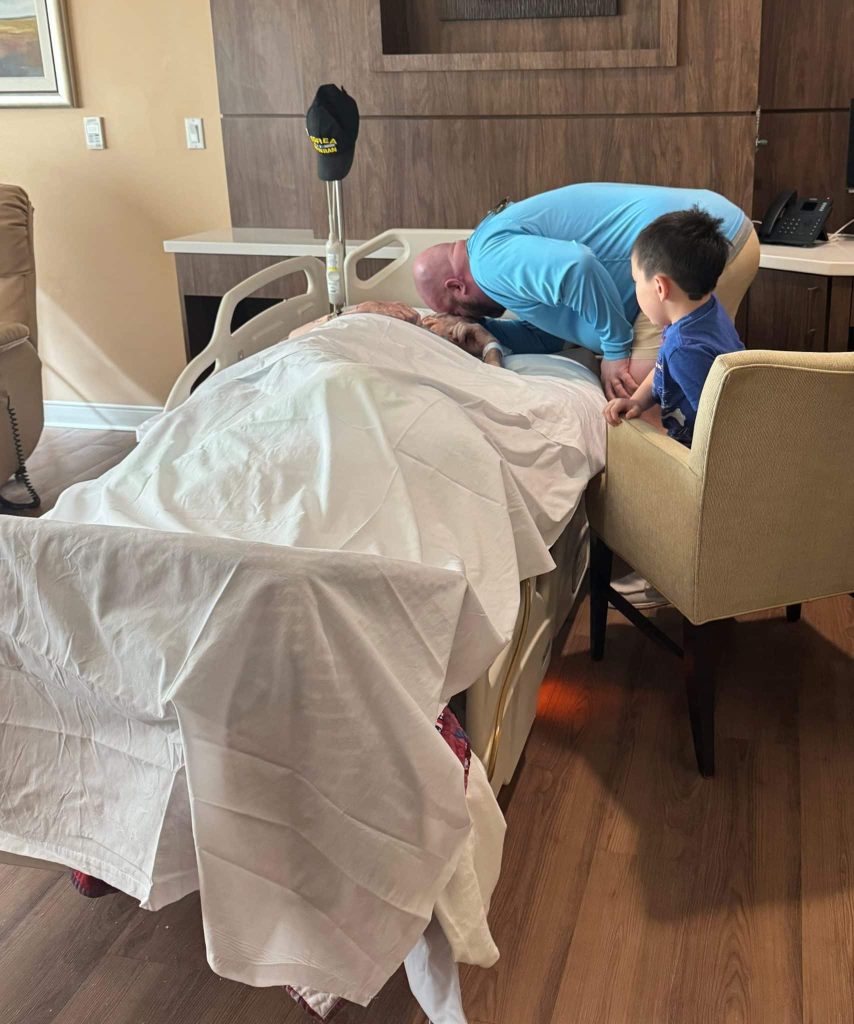 A son leaning over his father’s hospice bed with a grandson beside them, the father’s Korean War veteran cap hanging on the bed rail