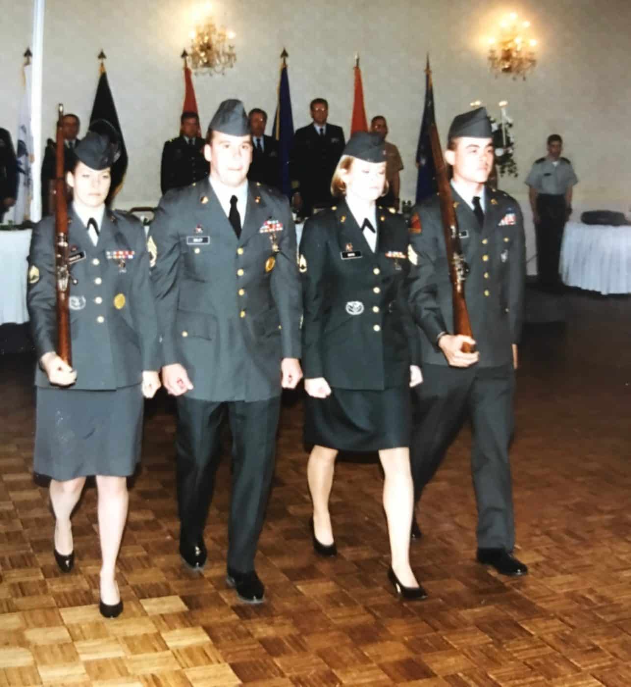 Four soldiers in dress uniform marching with rifles past a line of service flags