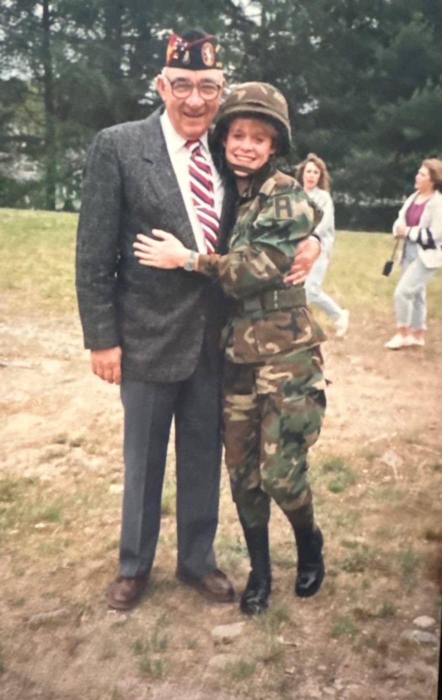 An older veteran in a service cap with his arm around a young soldier in camouflage uniform and helmet